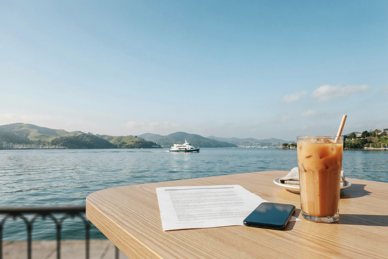 A printed document, a phone and an iced milk tea on a café table by the harbour, with a ferry and green hills behind