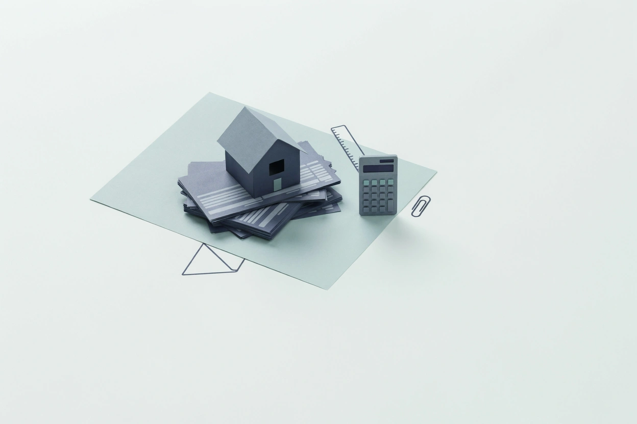 A small paper model house resting on a stack of pay stubs beside a calculator