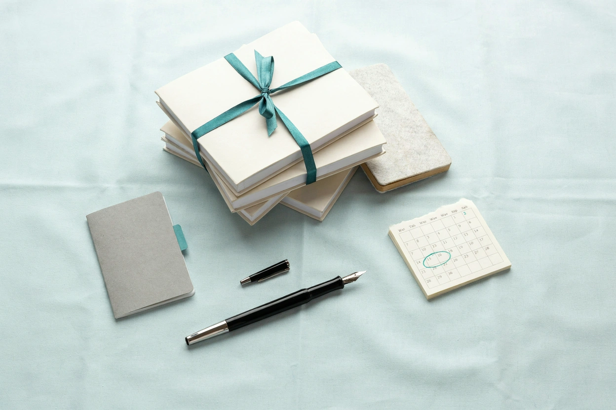 A stack of notebooks tied with ribbon beside a fountain pen and a calendar page with one date circled